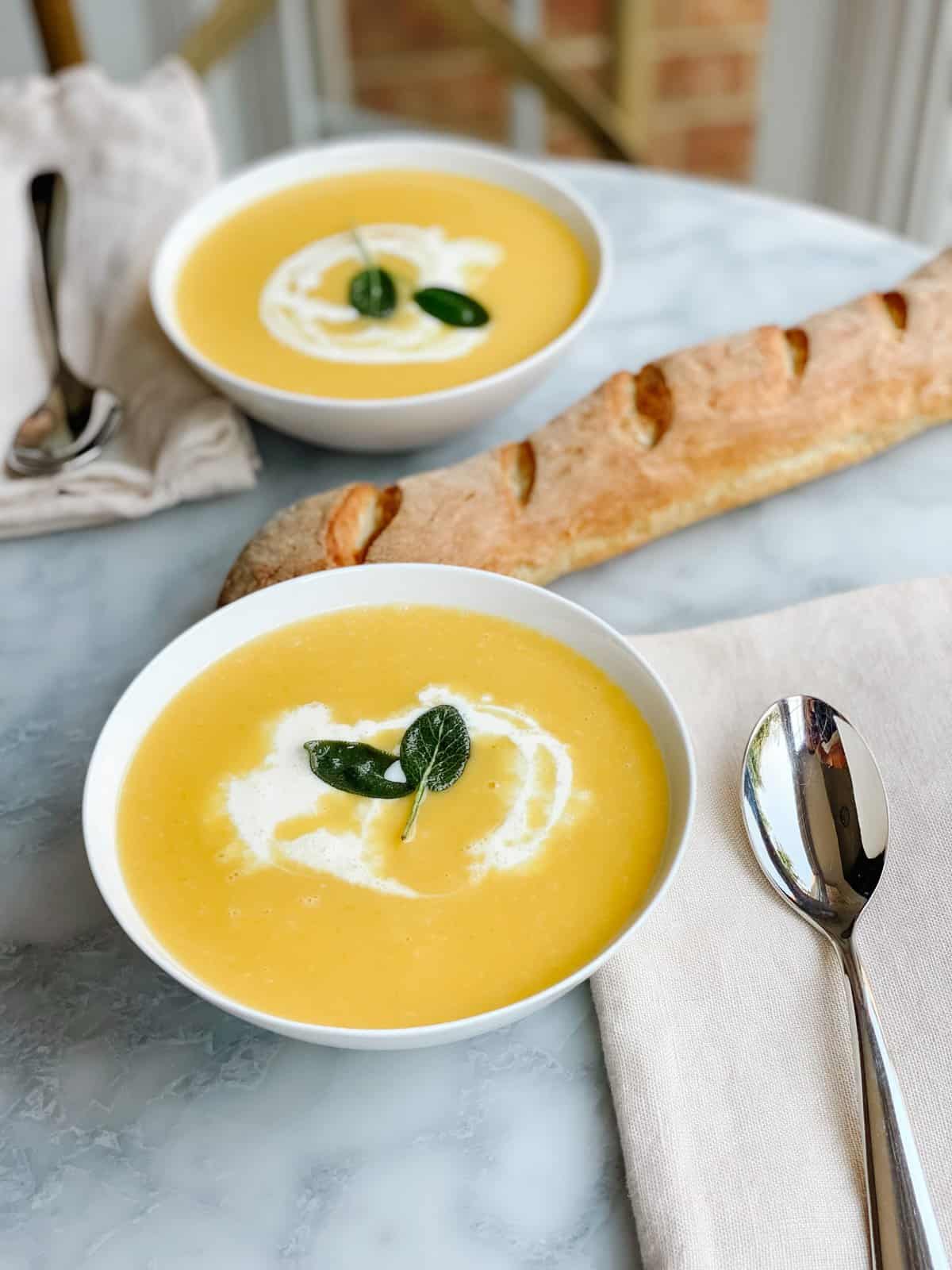 A white bowl of silky butternut squash soup swirled with cream and garnished with crispy sage leaves and a drizzle of olive oil on a wooden table