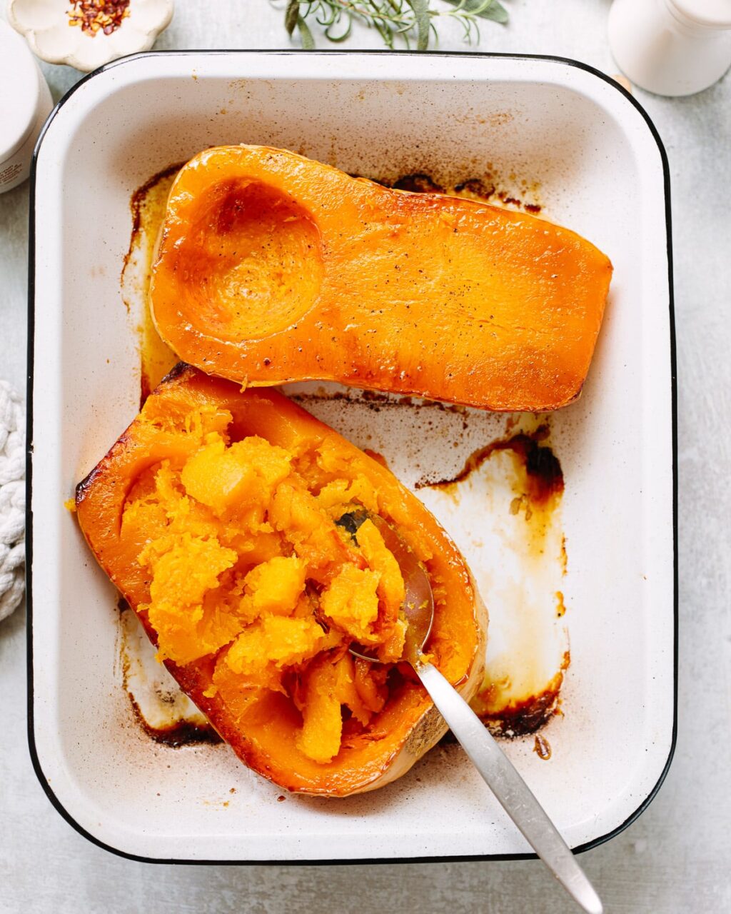 Halved butternut squash cut-side down on a baking sheet next to bowls of broth, sage leaves, garlic, and cream on a marble counter