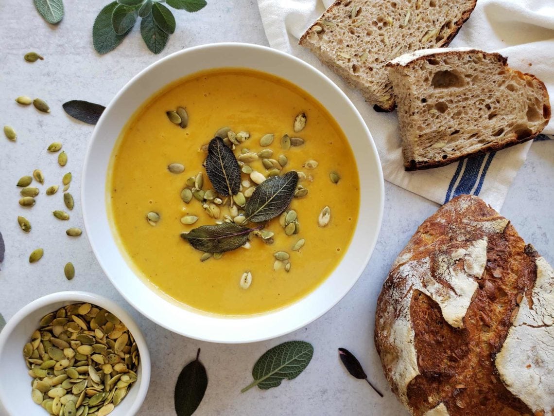 Close-up of a bowl of butternut squash soup with a swirl of cream, three crispy fried sage leaves, and a sprinkle of cracked black pepper, with a piece of crusty bread resting on the bowl edge