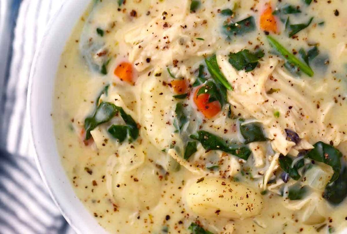 A bowl of creamy chicken gnocchi soup topped with fresh spinach and shredded carrots, served with crusty bread on a rustic wooden table