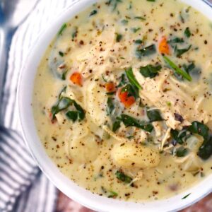 A bowl of creamy chicken gnocchi soup topped with fresh spinach and shredded carrots, served with crusty bread on a rustic wooden table