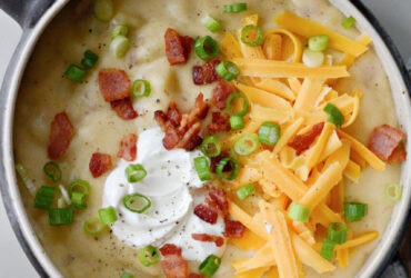 A rustic bowl of loaded baked potato soup topped with crispy bacon crumbles, shredded cheddar cheese, fresh chives, and a dollop of sour cream, styled on a wooden table with a linen napkin