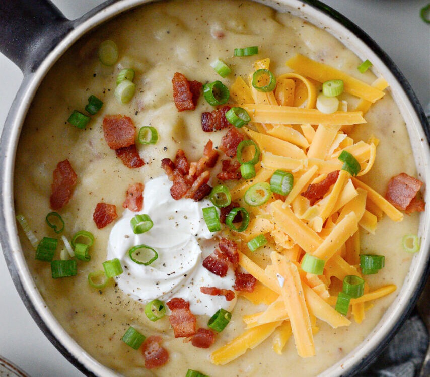 A rustic bowl of loaded baked potato soup topped with crispy bacon crumbles, shredded cheddar cheese, fresh chives, and a dollop of sour cream, styled on a wooden table with a linen napkin