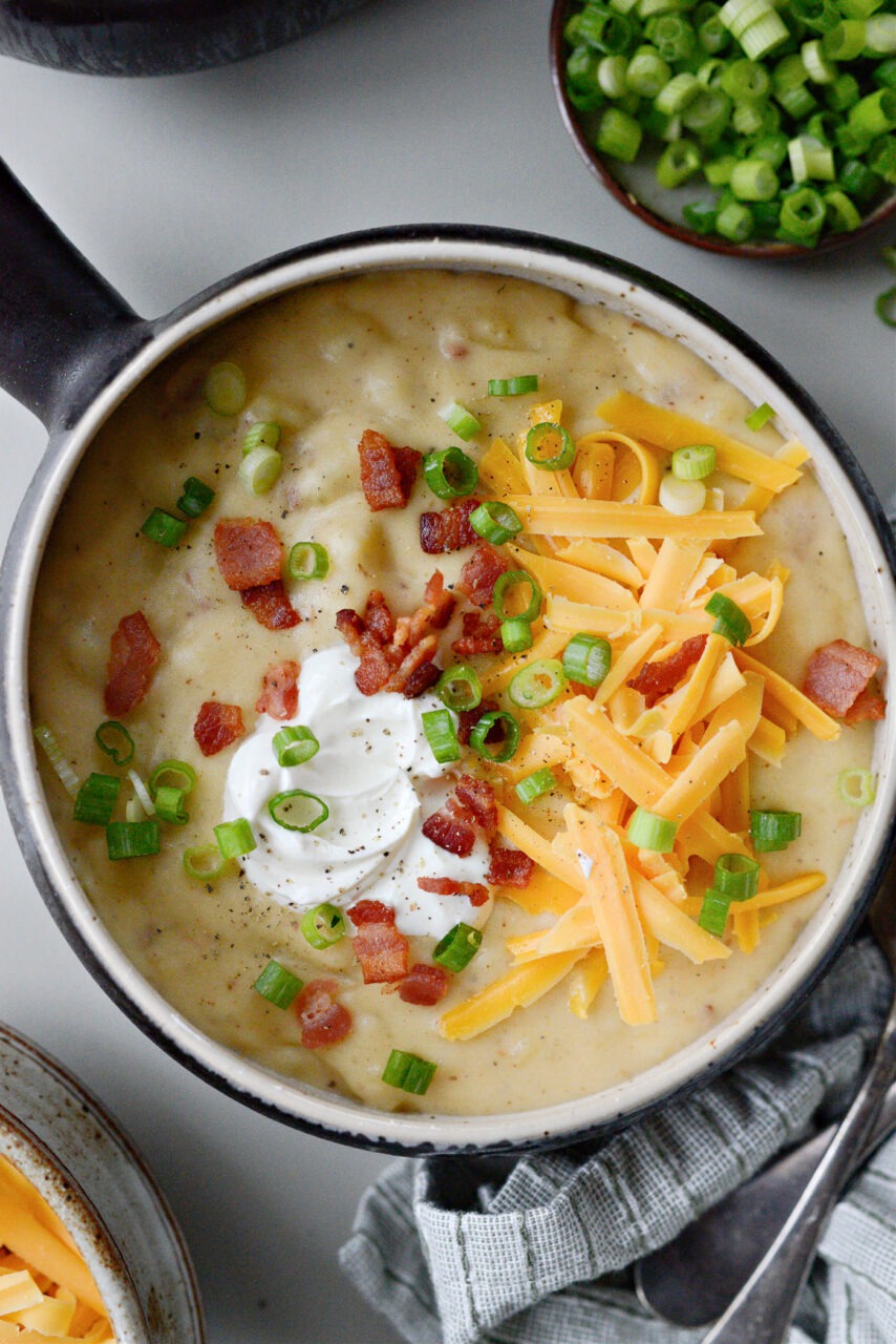 A rustic bowl of loaded baked potato soup topped with crispy bacon crumbles, shredded cheddar cheese, fresh chives, and a dollop of sour cream, styled on a wooden table with a linen napkin