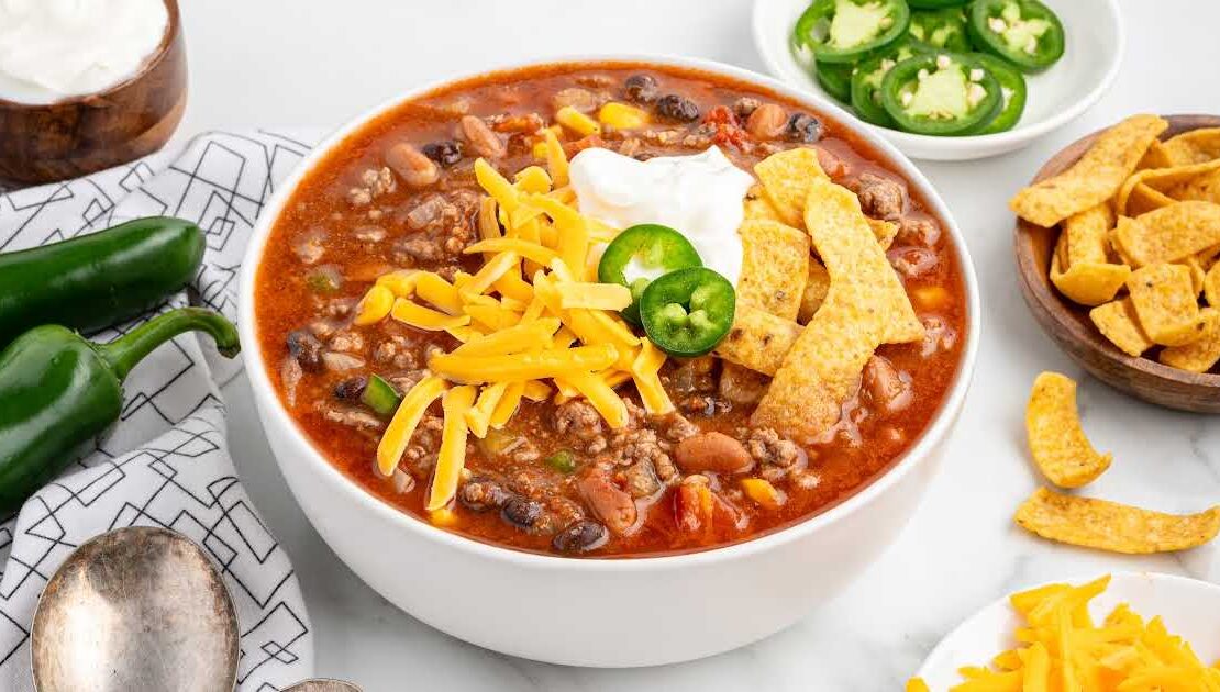 A rustic bowl of taco soup topped with shredded cheddar cheese, sour cream, sliced jalapeños, and crushed tortilla chips, photographed overhead on a dark wood table with colorful napkins