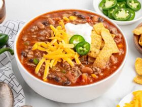 A rustic bowl of taco soup topped with shredded cheddar cheese, sour cream, sliced jalapeños, and crushed tortilla chips, photographed overhead on a dark wood table with colorful napkins