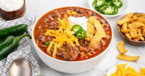 A rustic bowl of taco soup topped with shredded cheddar cheese, sour cream, sliced jalapeños, and crushed tortilla chips, photographed overhead on a dark wood table with colorful napkins