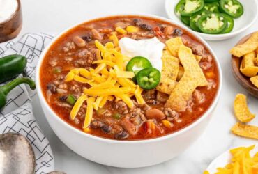 A rustic bowl of taco soup topped with shredded cheddar cheese, sour cream, sliced jalapeños, and crushed tortilla chips, photographed overhead on a dark wood table with colorful napkins