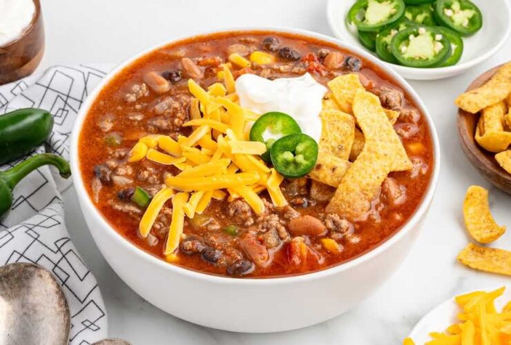 A rustic bowl of taco soup topped with shredded cheddar cheese, sour cream, sliced jalapeños, and crushed tortilla chips, photographed overhead on a dark wood table with colorful napkins
