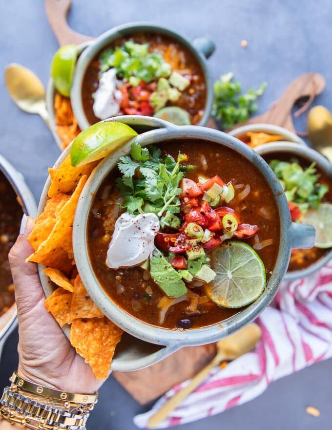 A close-up shot of a bowl of taco soup being garnished with a swirl of sour cream, a handful of shredded cheddar, and a sprinkle of fresh cilantro, with tortilla chips arranged on the side