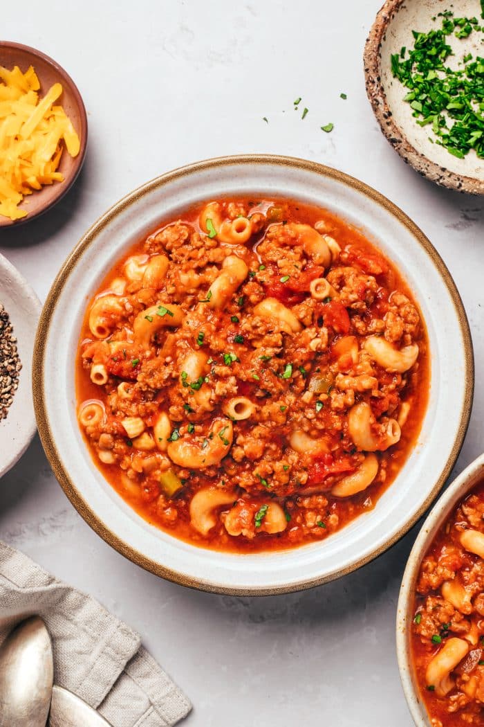 Overhead shot of ingredients for American goulash laid out on a kitchen counter — ground beef, elbow macaroni, canned tomatoes, broth, garlic, and spices