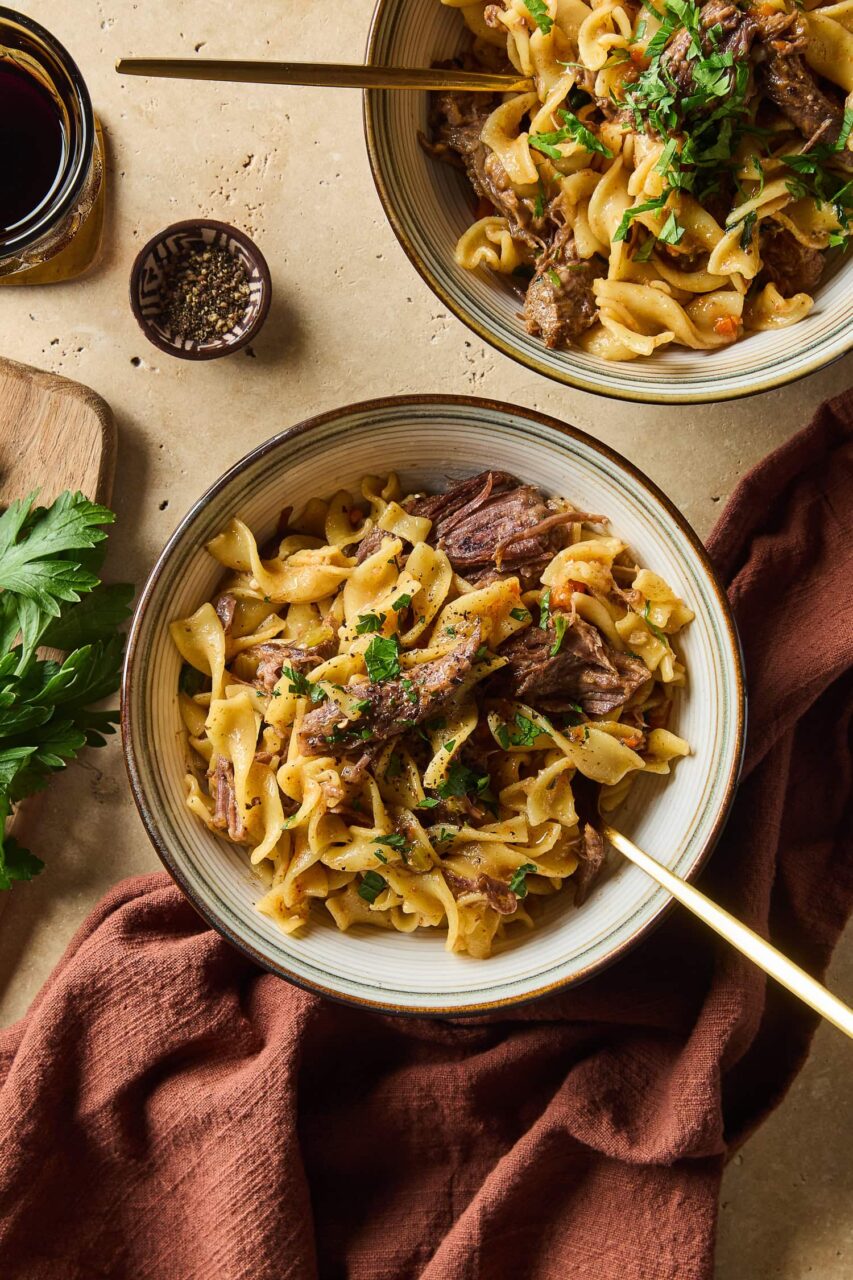 A bowl of beef and noodles with shredded beef over wide egg noodles in a rich brown gravy, garnished with fresh parsley