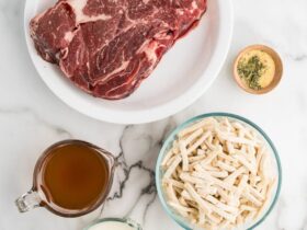 Overhead shot of ingredients for beef and noodles laid out on a wooden surface — chuck roast, egg noodles, beef broth, onion, garlic, and seasonings