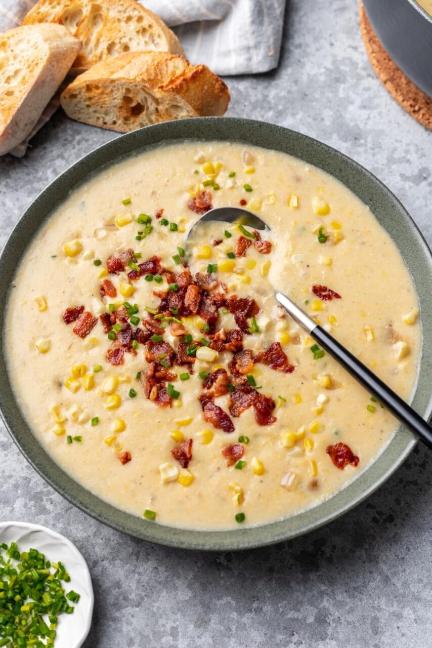 A rustic bowl of creamy corn chowder topped with crispy bacon crumbles, fresh chives, and a swirl of cream, photographed overhead on a wooden table
