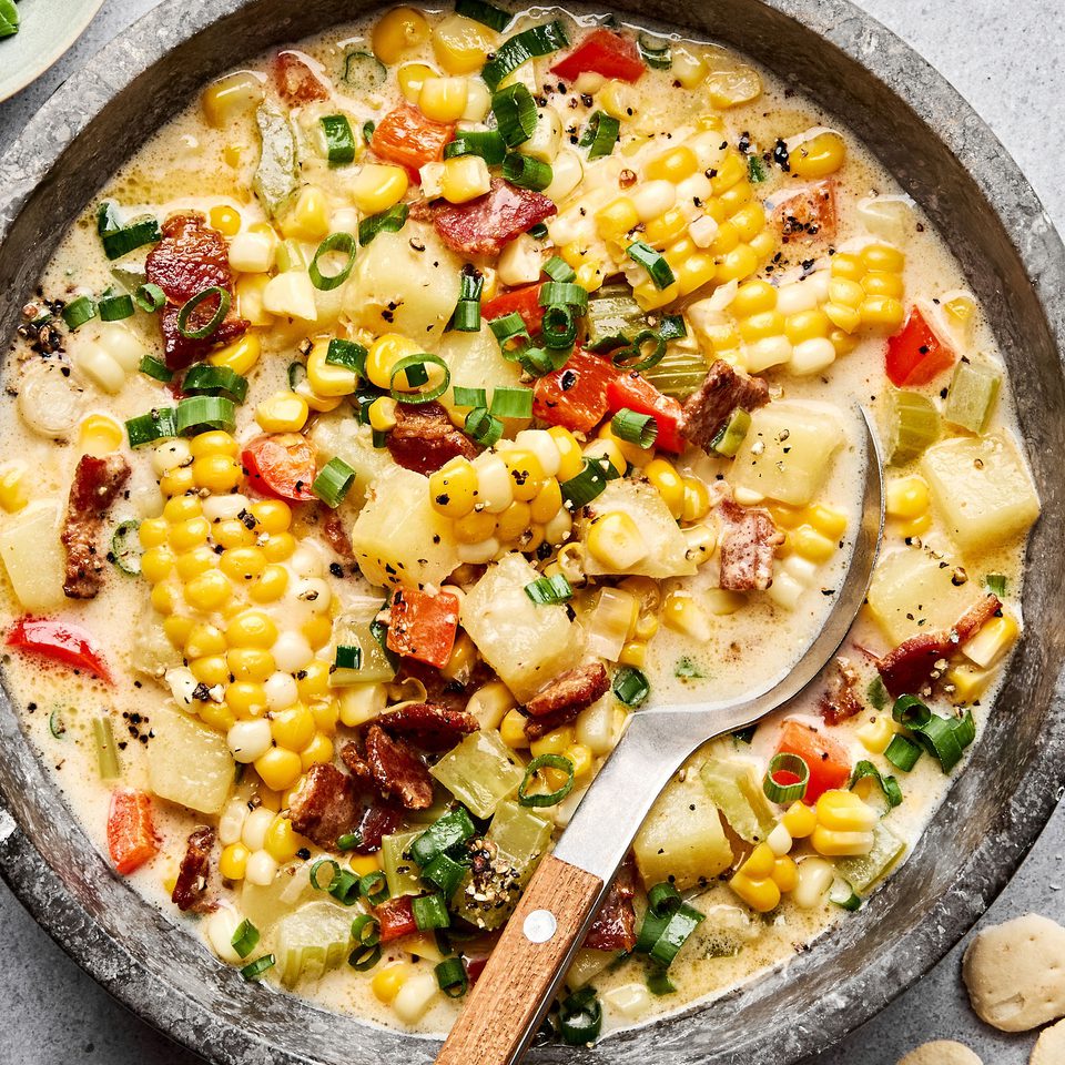 Side-by-side shot of raw corn chowder ingredients — diced potatoes, corn kernels, chopped bacon, onion, garlic, and cream — arranged on a cutting board next to a large pot