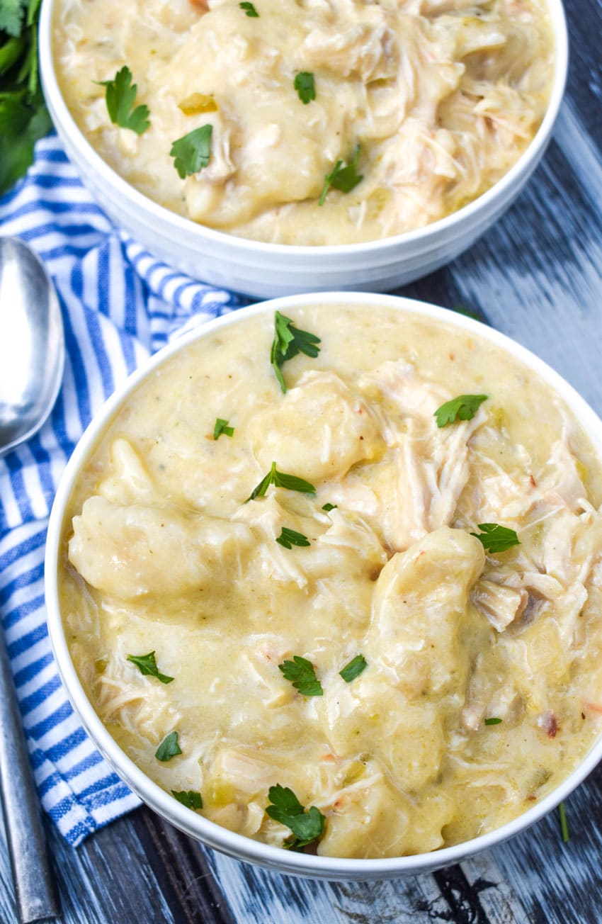 Crockpot chicken and dumplings served in a rustic white bowl with fluffy drop biscuit dumplings and creamy shredded chicken broth, overhead shot on a wooden farmhouse table