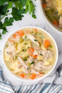 Flat lay of crockpot chicken and dumplings ingredients including raw chicken thighs, flour, cream of chicken soup cans, frozen vegetables, broth carton, butter, and fresh herbs on a light marble surface