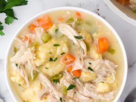 Flat lay of crockpot chicken and dumplings ingredients including raw chicken thighs, flour, cream of chicken soup cans, frozen vegetables, broth carton, butter, and fresh herbs on a light marble surface