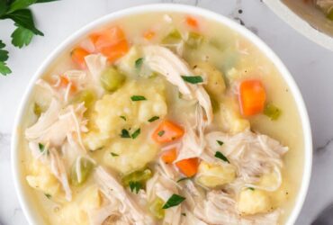 Flat lay of crockpot chicken and dumplings ingredients including raw chicken thighs, flour, cream of chicken soup cans, frozen vegetables, broth carton, butter, and fresh herbs on a light marble surface