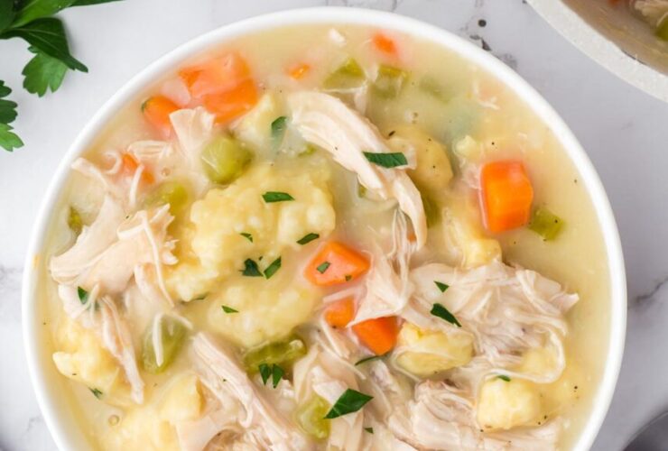 Flat lay of crockpot chicken and dumplings ingredients including raw chicken thighs, flour, cream of chicken soup cans, frozen vegetables, broth carton, butter, and fresh herbs on a light marble surface