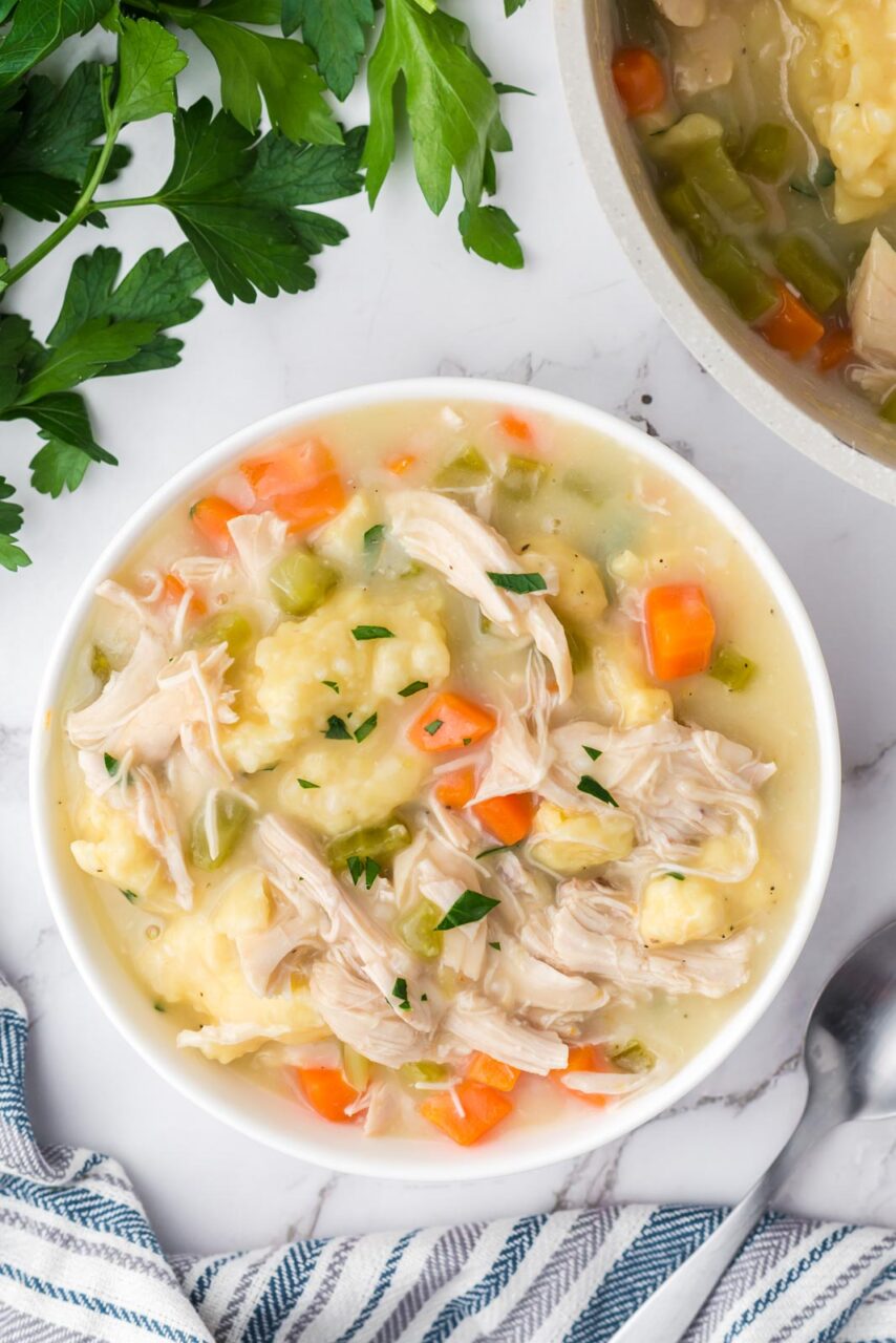 Flat lay of crockpot chicken and dumplings ingredients including raw chicken thighs, flour, cream of chicken soup cans, frozen vegetables, broth carton, butter, and fresh herbs on a light marble surface