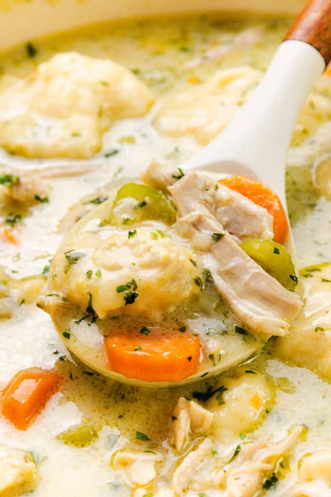 Close-up of a spoon lifting a fluffy drop biscuit dumpling from a bowl of creamy crockpot chicken and dumplings, steam rising, rustic farmhouse setting