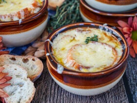 Step-by-step shot of caramelized onions cooking in a wide Dutch oven alongside ingredients: baguette slices, Gruyère cheese, beef broth, and fresh thyme on a kitchen counter