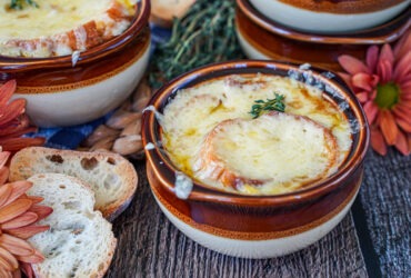 Step-by-step shot of caramelized onions cooking in a wide Dutch oven alongside ingredients: baguette slices, Gruyère cheese, beef broth, and fresh thyme on a kitchen counter