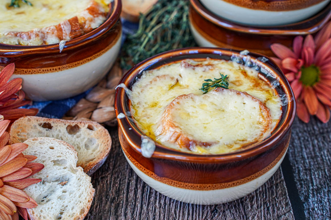 Step-by-step shot of caramelized onions cooking in a wide Dutch oven alongside ingredients: baguette slices, Gruyère cheese, beef broth, and fresh thyme on a kitchen counter