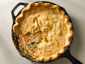 Golden flaky turkey pot pie in a cast iron skillet with a perfectly crimped crust and steam venting through slits, shot on a rustic wooden table with a serving spoon and golden-brown filling visible through a cut slice