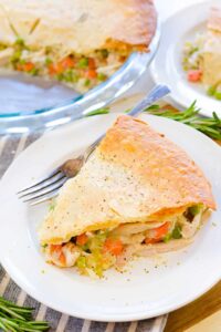 Close-up of a serving of turkey pot pie on a white plate showing the creamy golden filling with chunks of turkey, peas, carrots and corn spilling out from beneath the flaky, buttery top crust