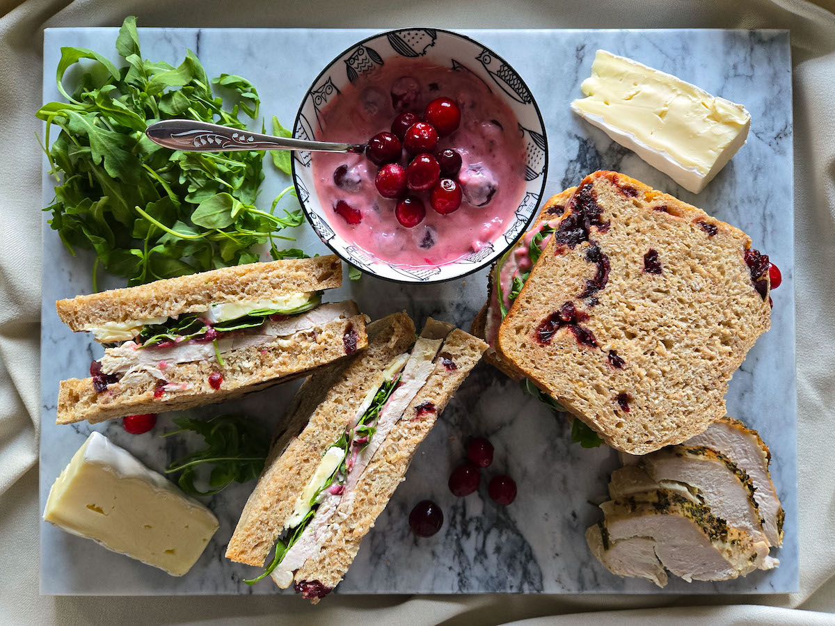 Overhead flat lay of leftover turkey sandwich ingredients — pulled turkey, cranberry sauce, mayonnaise, stuffing, arugula, and sourdough bread on a marble countertop