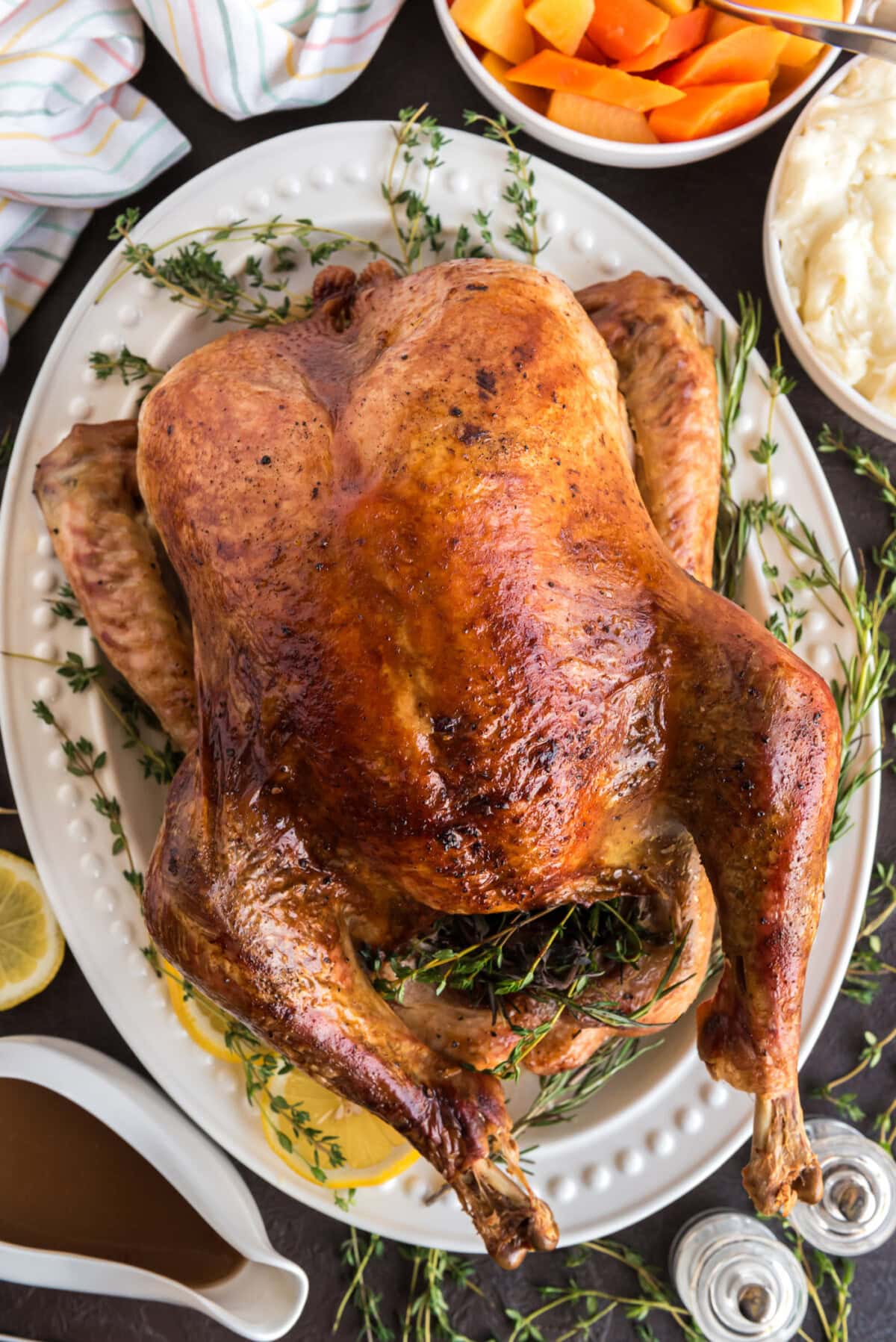 A close-up of a beautifully carved Thanksgiving turkey on a serving platter surrounded by fresh herb garnishes, with bowls of cranberry sauce, stuffing, and gravy in soft focus in the background