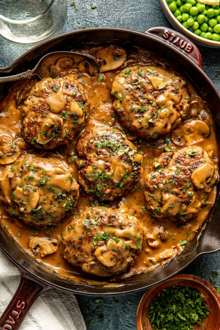 Raw Salisbury steak beef patties with mushrooms, onion, Worcestershire sauce, breadcrumbs, egg, and beef broth ingredients laid out on a wooden cutting board