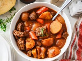 Flat lay of slow cooker beef stew ingredients including cubed chuck roast, diced carrots, Yukon Gold potatoes, garlic cloves, tomato paste, Worcestershire sauce, beef broth, and dried herbs on a wooden cutting board
