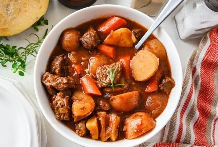 Flat lay of slow cooker beef stew ingredients including cubed chuck roast, diced carrots, Yukon Gold potatoes, garlic cloves, tomato paste, Worcestershire sauce, beef broth, and dried herbs on a wooden cutting board