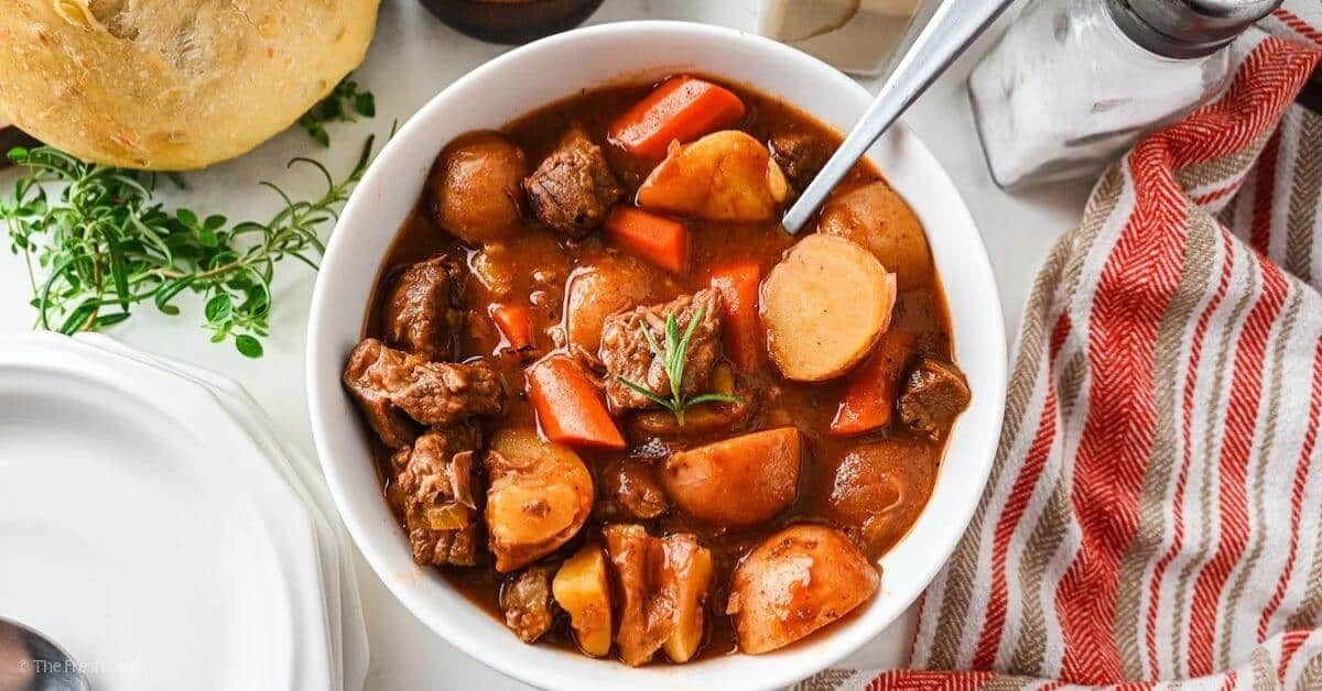 Flat lay of slow cooker beef stew ingredients including cubed chuck roast, diced carrots, Yukon Gold potatoes, garlic cloves, tomato paste, Worcestershire sauce, beef broth, and dried herbs on a wooden cutting board