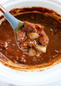 Close-up of a spoonful of slow cooker beef stew being lifted from a rustic bowl, showing glossy rich gravy coating fork-tender beef and soft potato chunks, with steam rising