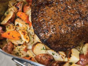 Chuck roast being seared in a cast iron skillet with golden brown crust forming, surrounded by measured spices, carrots, and Yukon Gold potatoes on a wood cutting board