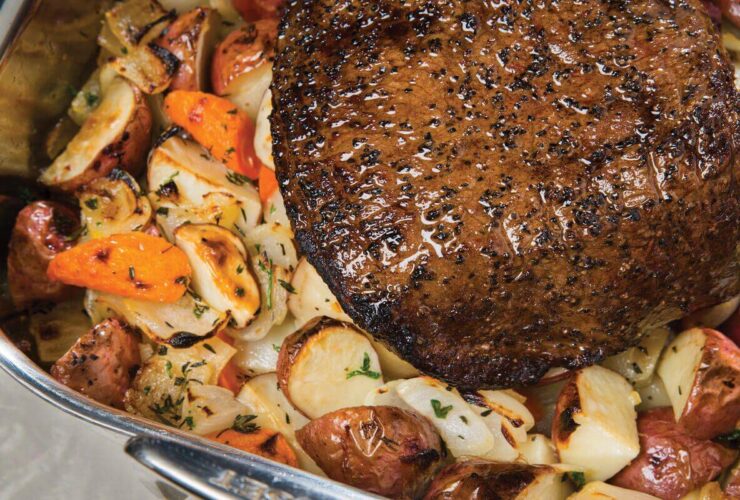 Chuck roast being seared in a cast iron skillet with golden brown crust forming, surrounded by measured spices, carrots, and Yukon Gold potatoes on a wood cutting board