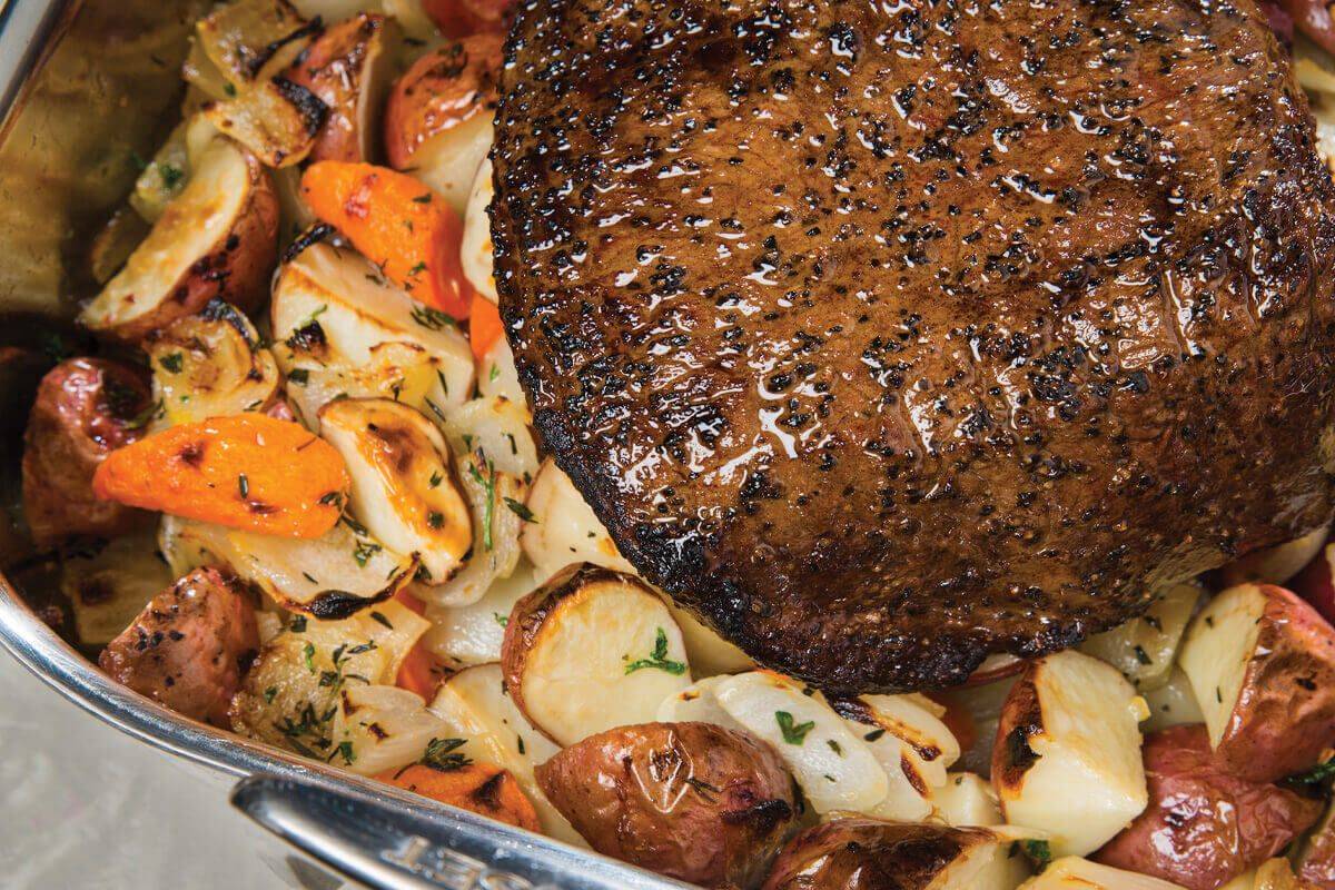 Chuck roast being seared in a cast iron skillet with golden brown crust forming, surrounded by measured spices, carrots, and Yukon Gold potatoes on a wood cutting board