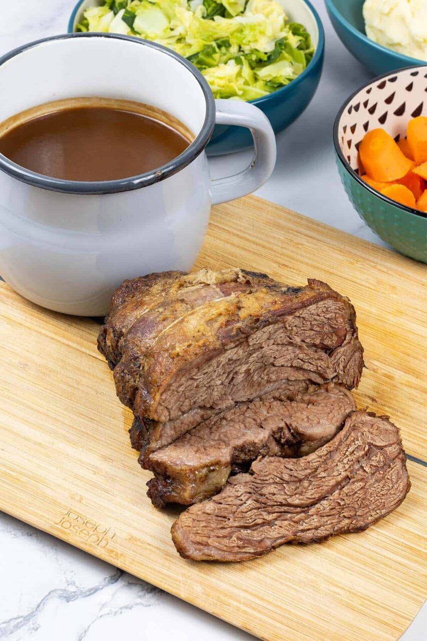 Close-up of slow cooker pot roast sliced on a wooden cutting board with glossy brown gravy being spooned over tender beef, fresh parsley garnish, with a fork resting beside it