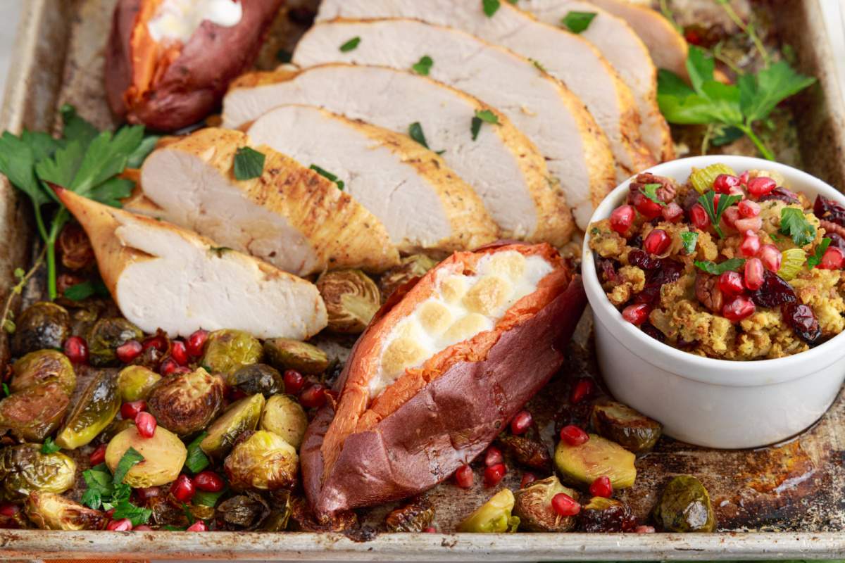 Overhead flat-lay of all the ingredients for a small Thanksgiving dinner including a turkey breast, fresh herbs, bread cubes, cranberries, green beans, and small bowls of butter and broth on a marble surface