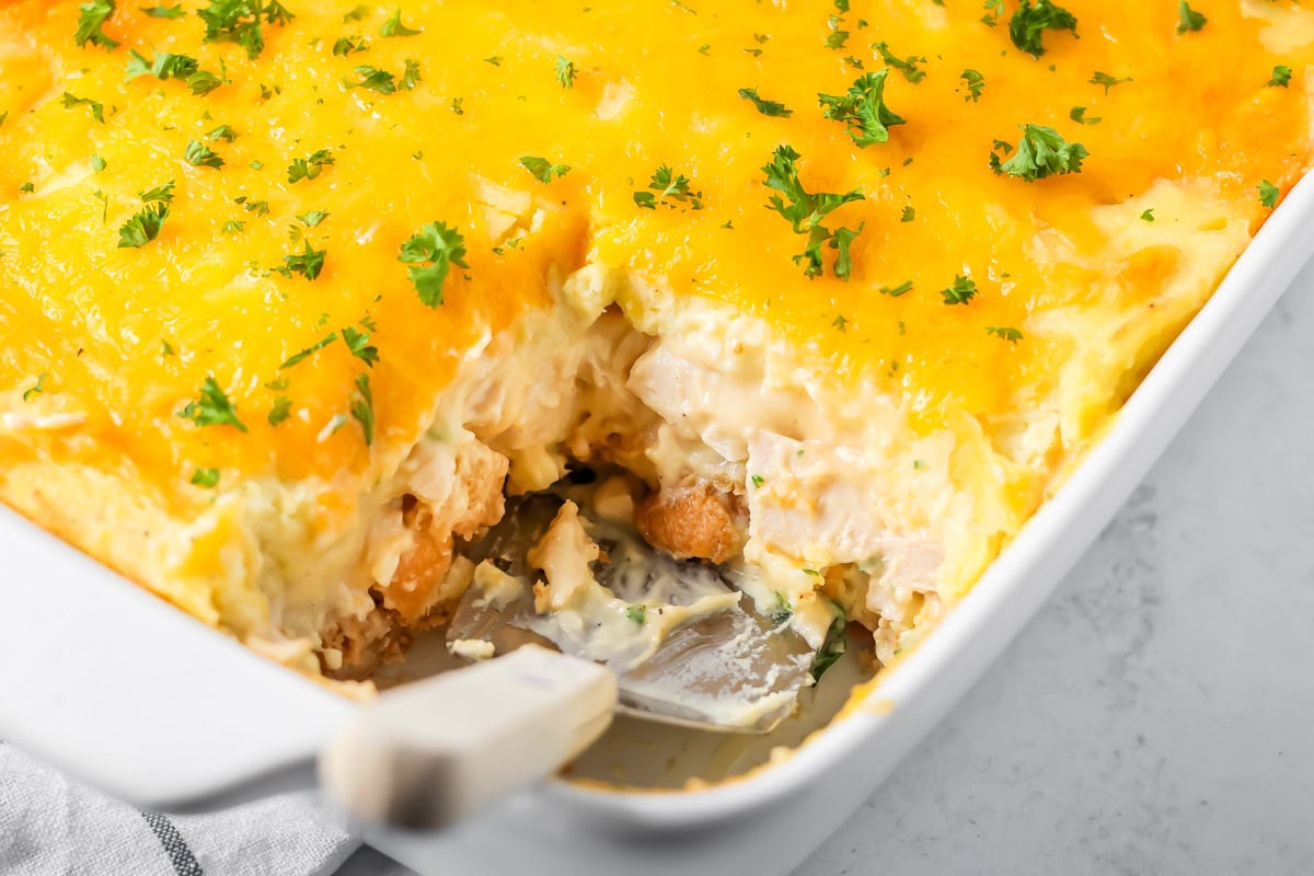 Golden-topped Thanksgiving leftover casserole in a white baking dish with layers of mashed potatoes, stuffing, and turkey visible at the edges, served on a wooden table with a sprig of fresh thyme