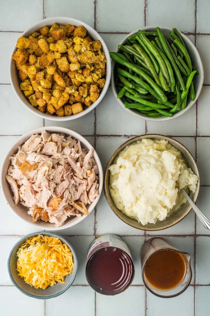 Overhead flat-lay of Thanksgiving leftover casserole ingredients — shredded turkey, stuffing, mashed potatoes, gravy, and shredded cheese arranged in separate bowls on a rustic surface