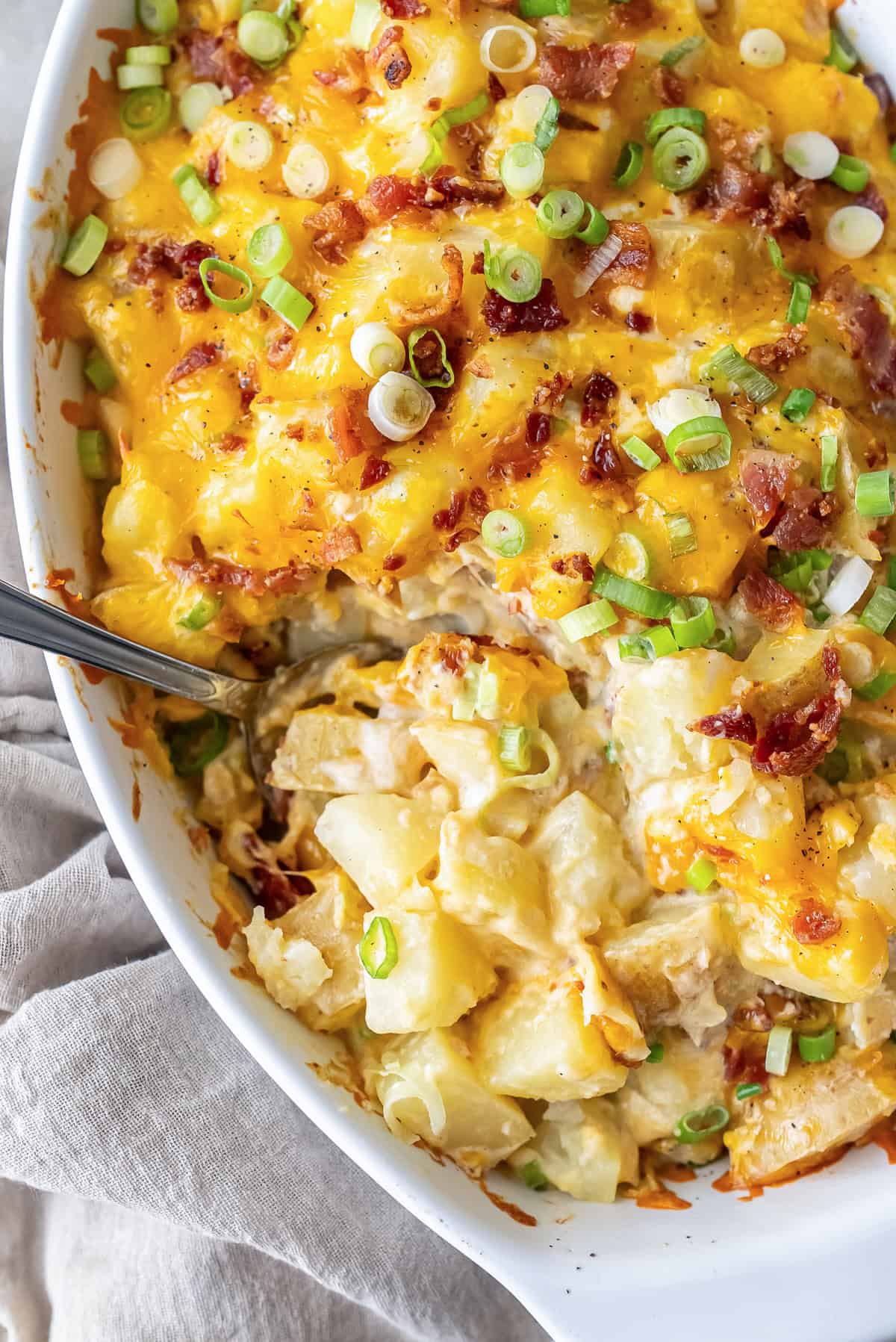 Twice baked potato casserole in a white 9x13 baking dish, bubbling with golden cheddar cheese, crispy bacon crumbles, and green onions on top, photographed from above on a rustic wooden table