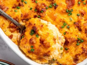 Overhead flat-lay of ingredients for twice baked potato casserole — russet potatoes, shredded cheddar, sour cream, cream cheese, bacon strips, green onions, butter, and garlic powder arranged on a light marble surface