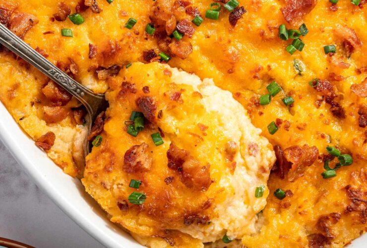 Overhead flat-lay of ingredients for twice baked potato casserole — russet potatoes, shredded cheddar, sour cream, cream cheese, bacon strips, green onions, butter, and garlic powder arranged on a light marble surface