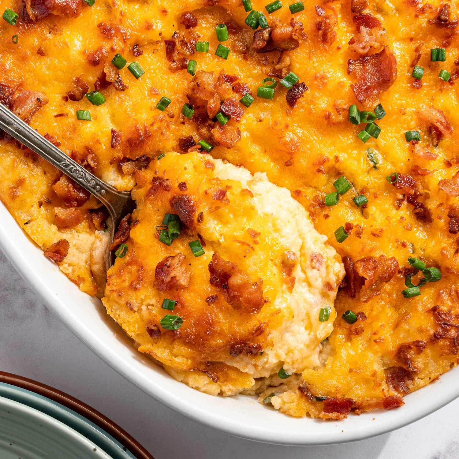 Overhead flat-lay of ingredients for twice baked potato casserole — russet potatoes, shredded cheddar, sour cream, cream cheese, bacon strips, green onions, butter, and garlic powder arranged on a light marble surface
