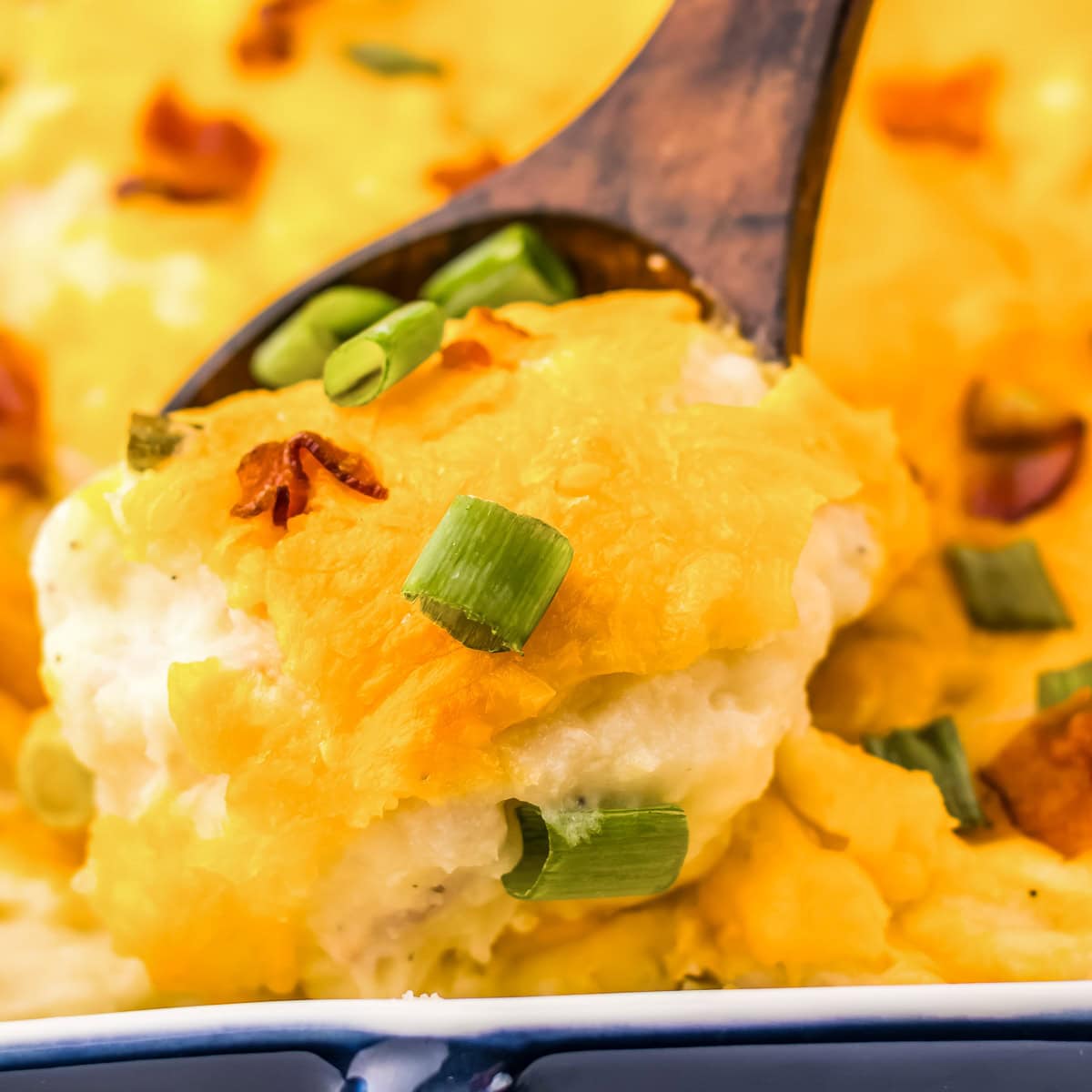 Close-up of a wooden spoon scooping a generous portion of twice baked potato casserole, showing the creamy interior with visible bacon bits, melted cheese pull, and green onion garnish